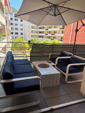 Un espace extérieur aménagé avec une table et deux canapés, un grand parasol et une vue sur un immeuble d’habitation.