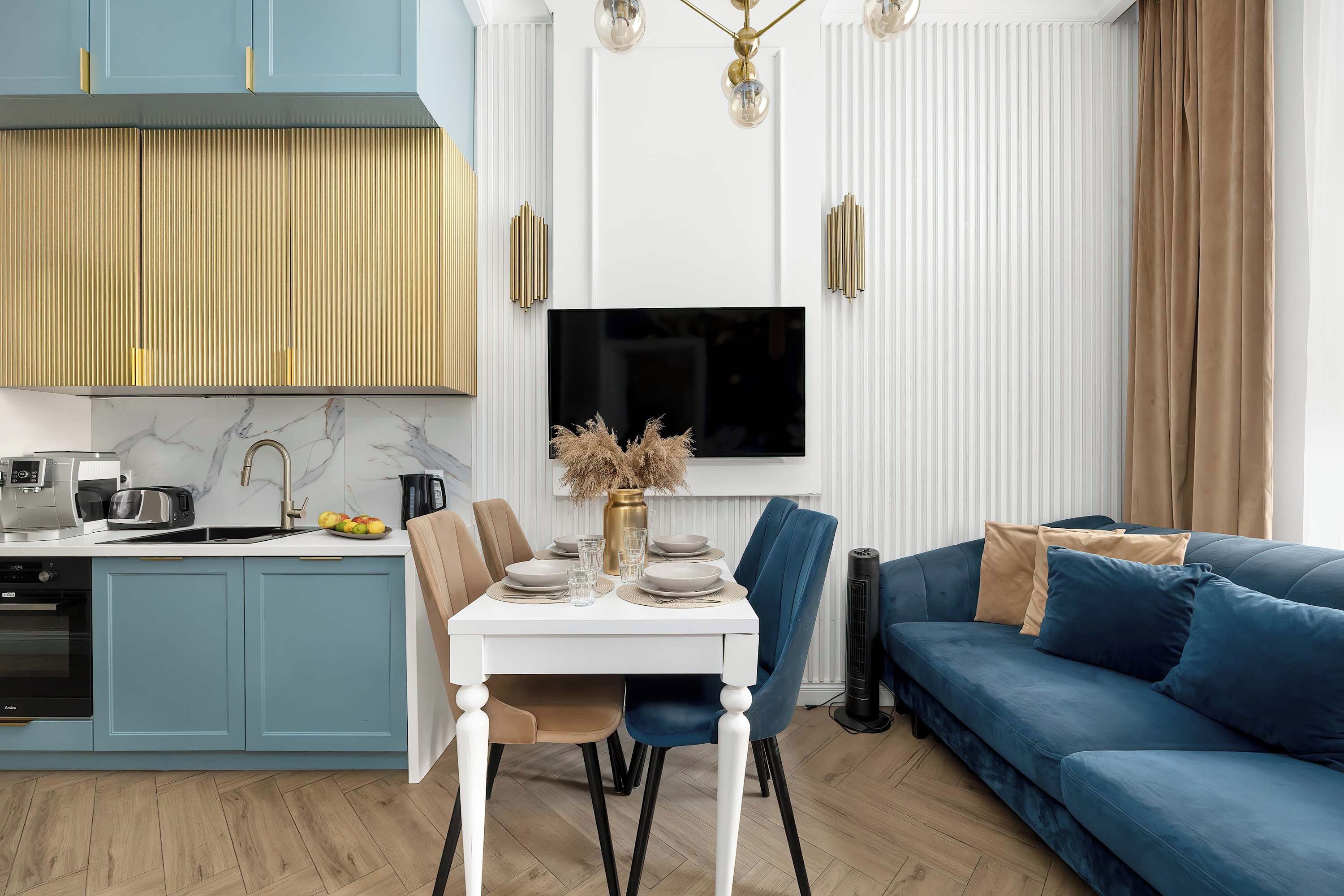 A modern kitchen with a built-in oven, a dining area with a white table and chairs, and a blue sofa in the living room.