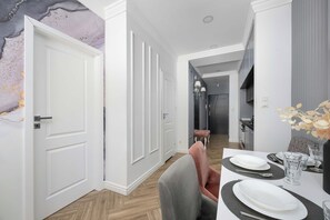 Ett modernt matområde med ett bord dukat för två personer, en hall med dörrar och ett kök som syns i bakgrunden.