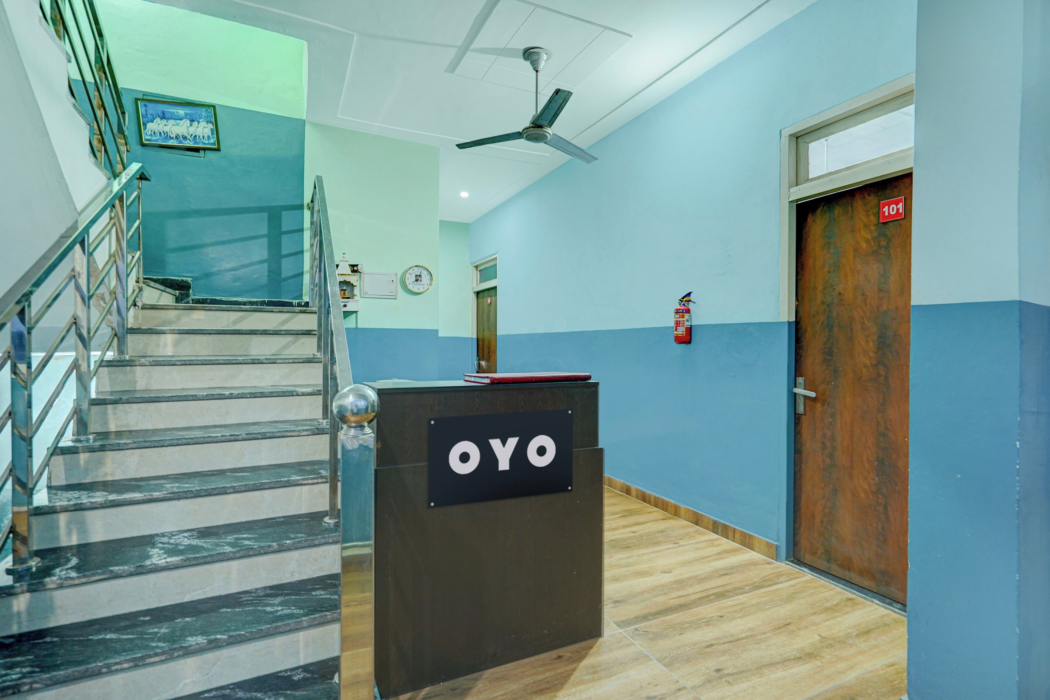 Un hall d’hôtel avec un comptoir d’accueil portant l’inscription « OYO », un escalier, un ventilateur de plafond et un extincteur.
