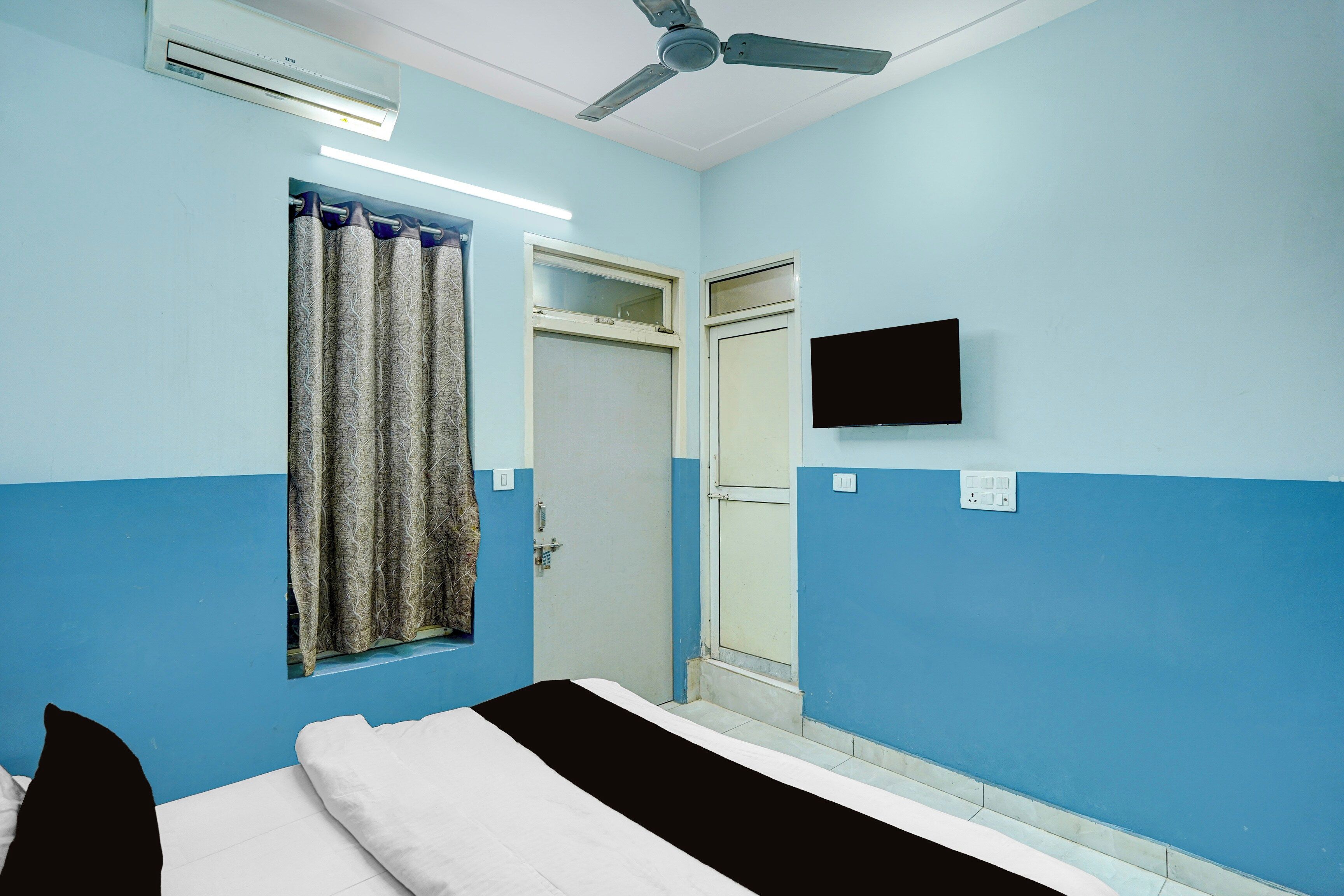 Une chambre avec un lit, un ventilateur de plafond, une fenêtre avec des rideaux et un téléviseur fixé au mur.