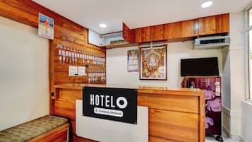 A hotel reception desk with a sign indicating it's a company-serviced hotel. There are wooden paneling on the walls, a bench, and a wall-mounted calendar.