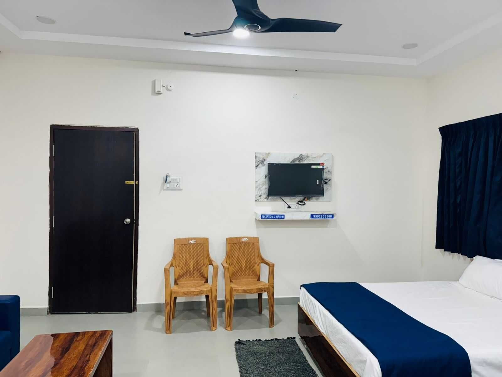 Habitación de hotel con cama, escritorio, dos sillas, televisor montado en la pared, ventilador de techo y una puerta.