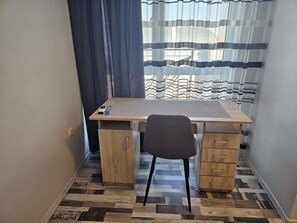 Ein kleiner, ordentlicher Arbeitsplatz mit Schreibtisch, Stuhl und Blick durch ein Fenster mit gestreifter Gardine.