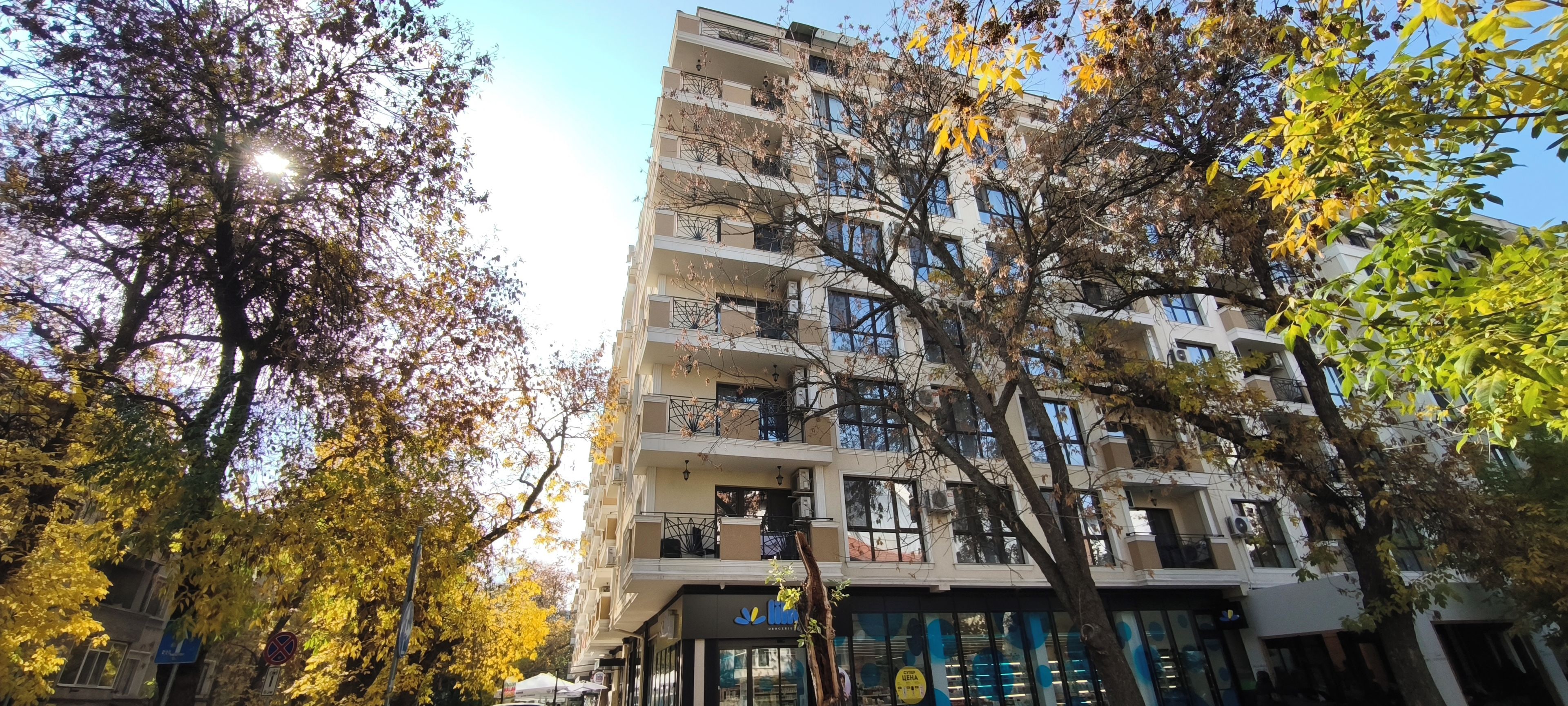 Un immeuble résidentiel à plusieurs étages, doté de balcons, avec un local commercial au rez-de-chaussée et des arbres aux feuilles d’automne.