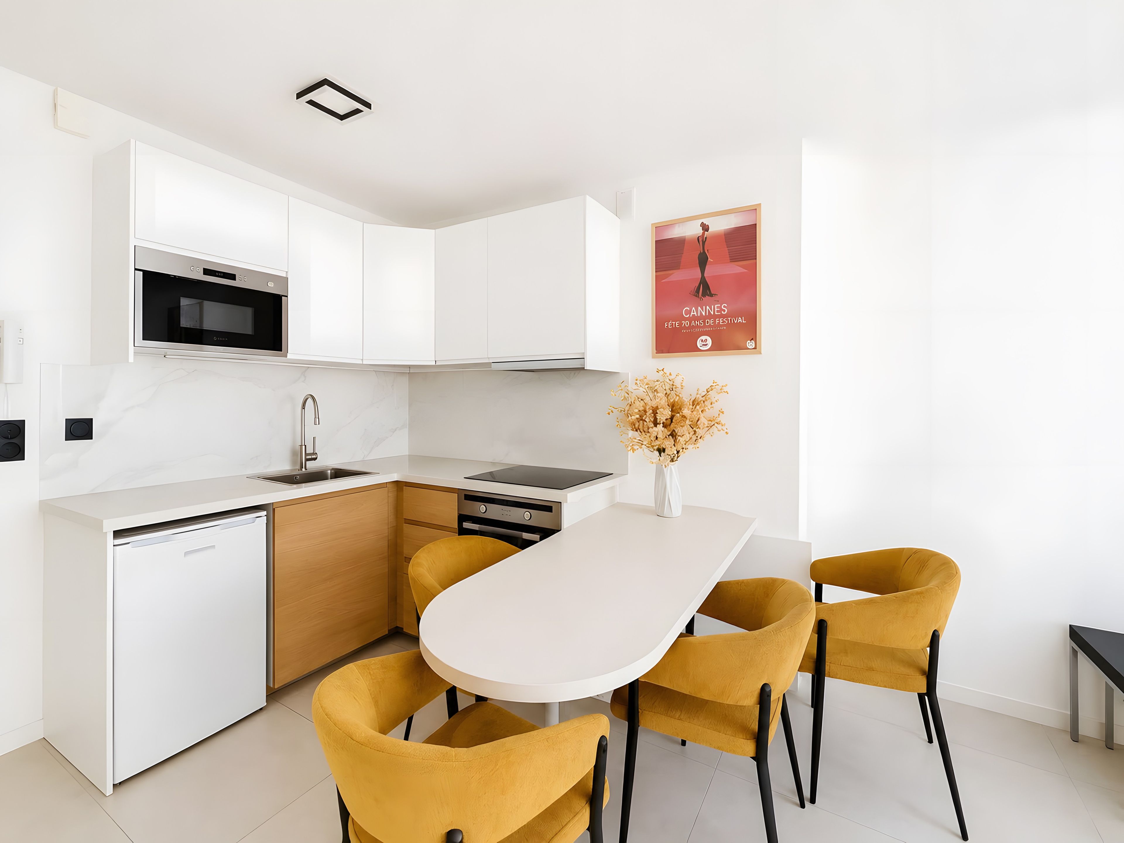 A modern kitchen with white cabinets, a built-in oven, and a dining area with a white table and mustard chairs.