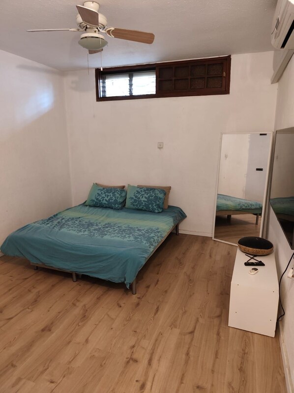 A bedroom with a bed, a ceiling fan, a window, a mirror, and a small white table with a basket.