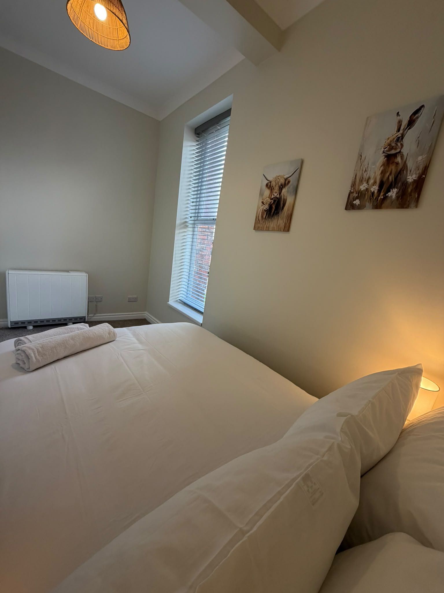 Un dormitorio con cama, un radiador, una ventana con persianas y dos cuadros colgados en la pared.