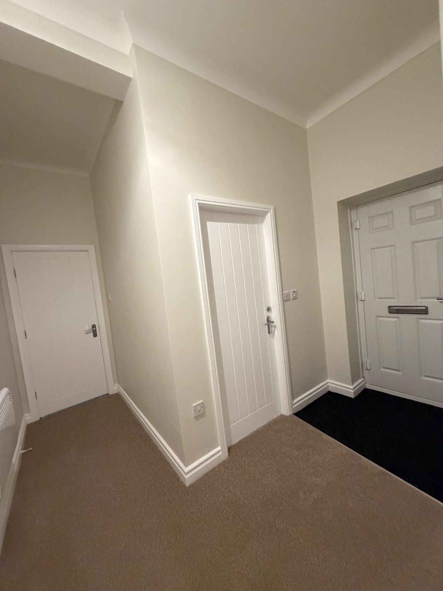Un pasillo con dos puertas blancas, alfombra color crema y una zona alfombrada de color negro que conduce a otra habitación.