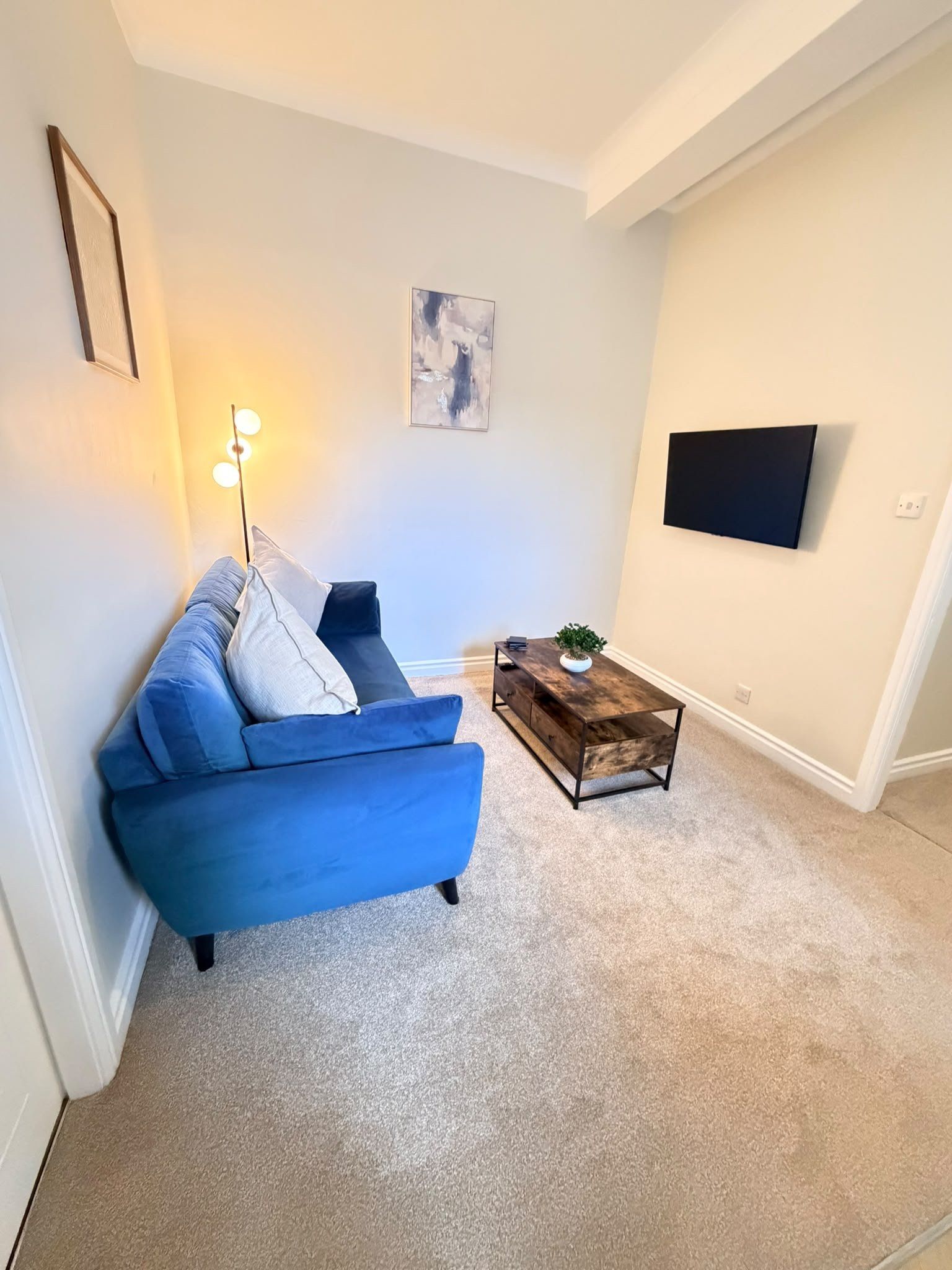 Una sala de estar con un sofá azul, una pequeña mesa de centro de madera, una lámpara de pie, un cuadro en la pared y un televisor de pantalla plana montado en la pared.