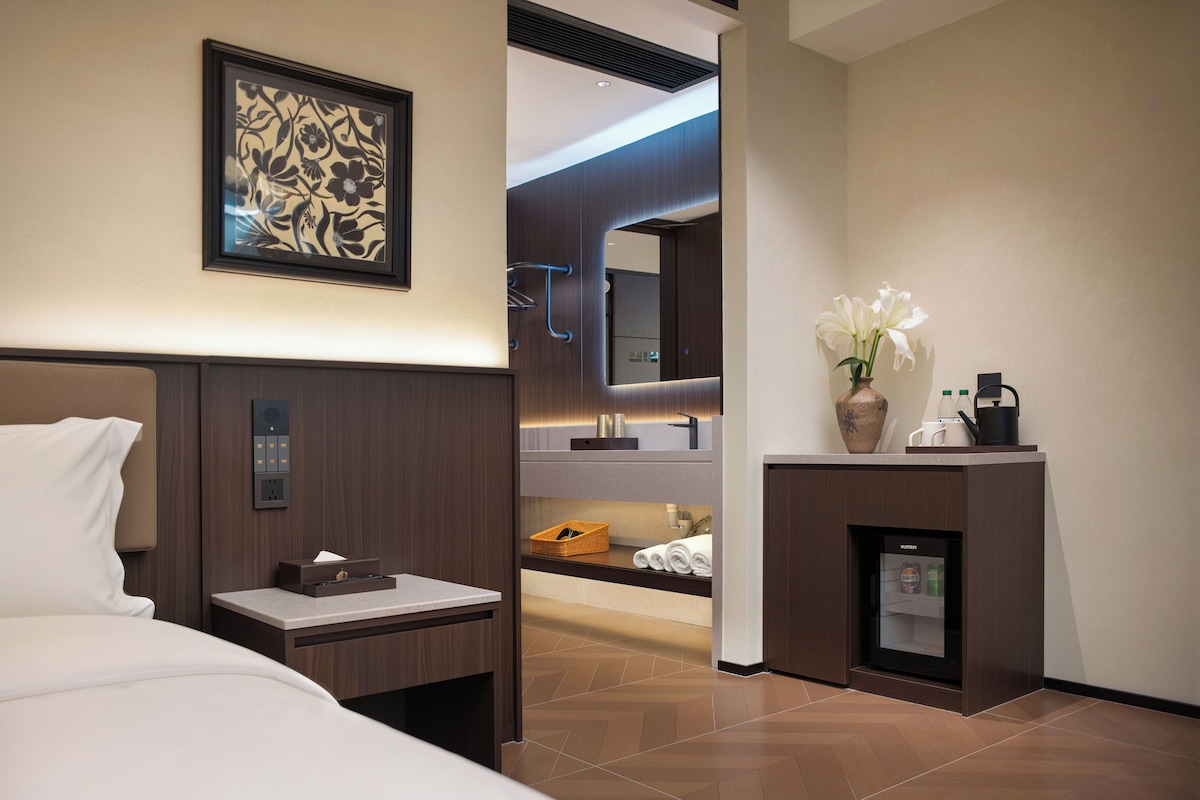 Een moderne hotelkamer met een bed, nachtkastje, een kleine koelkast, een spiegel en een kunstwerk in een lijst aan de muur.