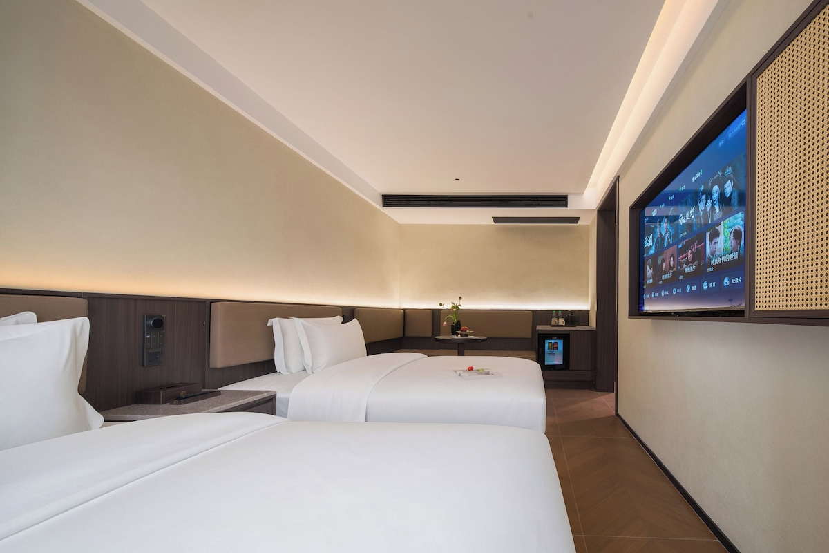 Een hotelkamer met twee bedden, een televisie, een klein tafeltje met een vaas en een wandgemonteerde beeldscherm.