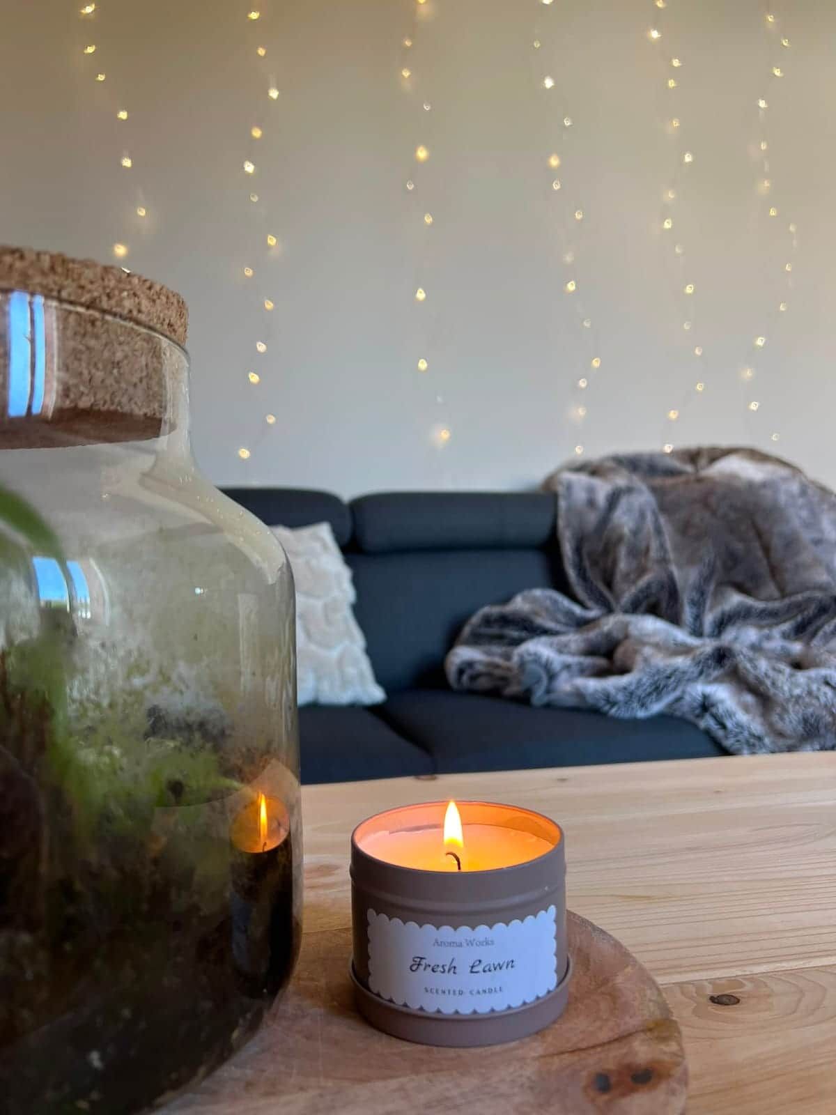A candle on a wooden coaster, a glass jar with a cork lid, and a dark-colored sofa with a blanket in a room with string lights.
