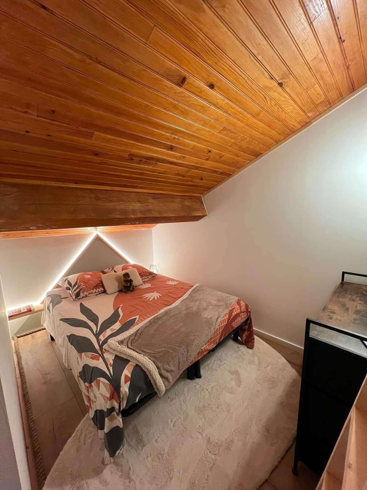 Une chambre avec un plafond en pente, un lit recouvert d’une courtepointe à motifs, une petite table et une chaise.