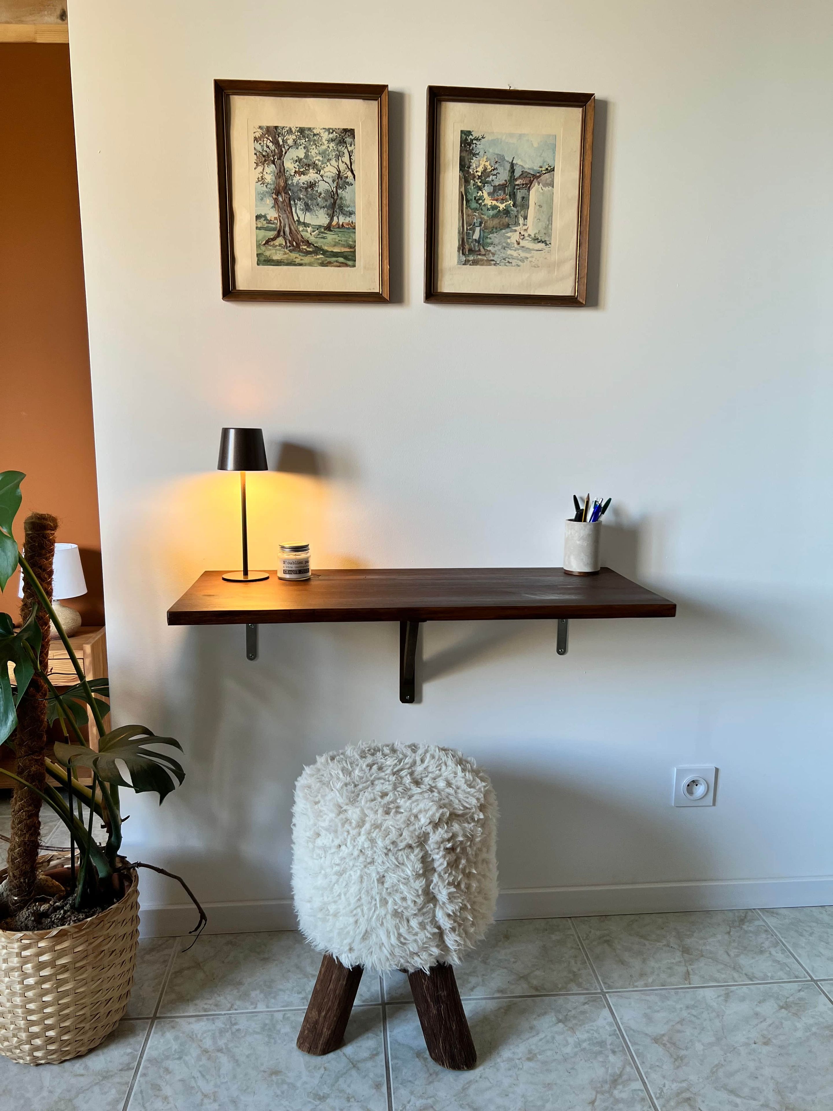 Une pièce dans laquelle on trouve une étagère en bois, sur laquelle sont posées deux œuvres d’art encadrées, une lampe et un petit tabouret.