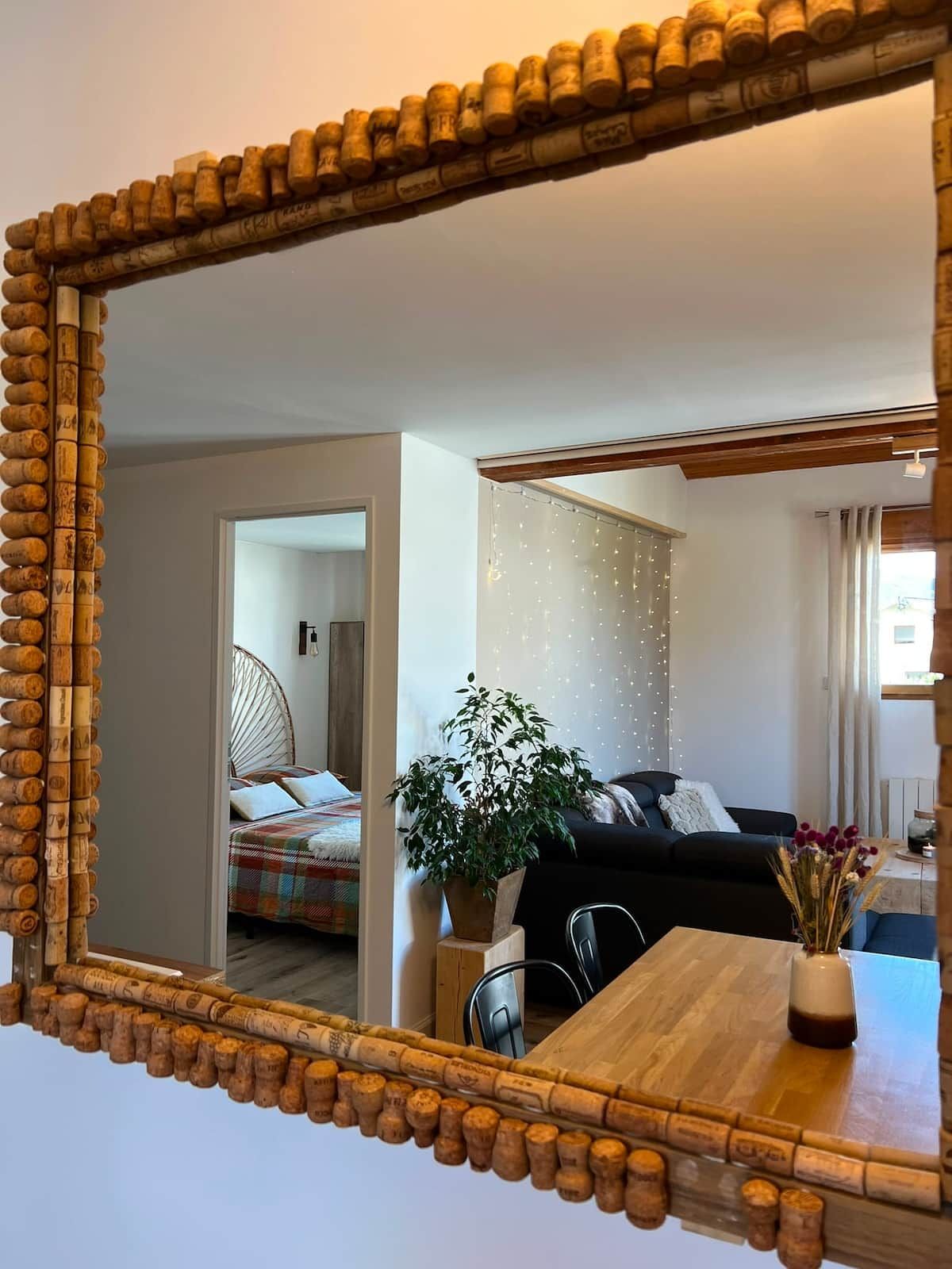 Ein Zimmer mit einem Holztisch, einem Spiegel mit Korkrahmen, einer Topfpflanze und einem schwarzen Sofa.
