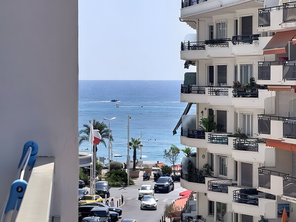 Vista de una calle costera con una vista despejada del mar, coches estacionados y un edificio de varios pisos con balcones.