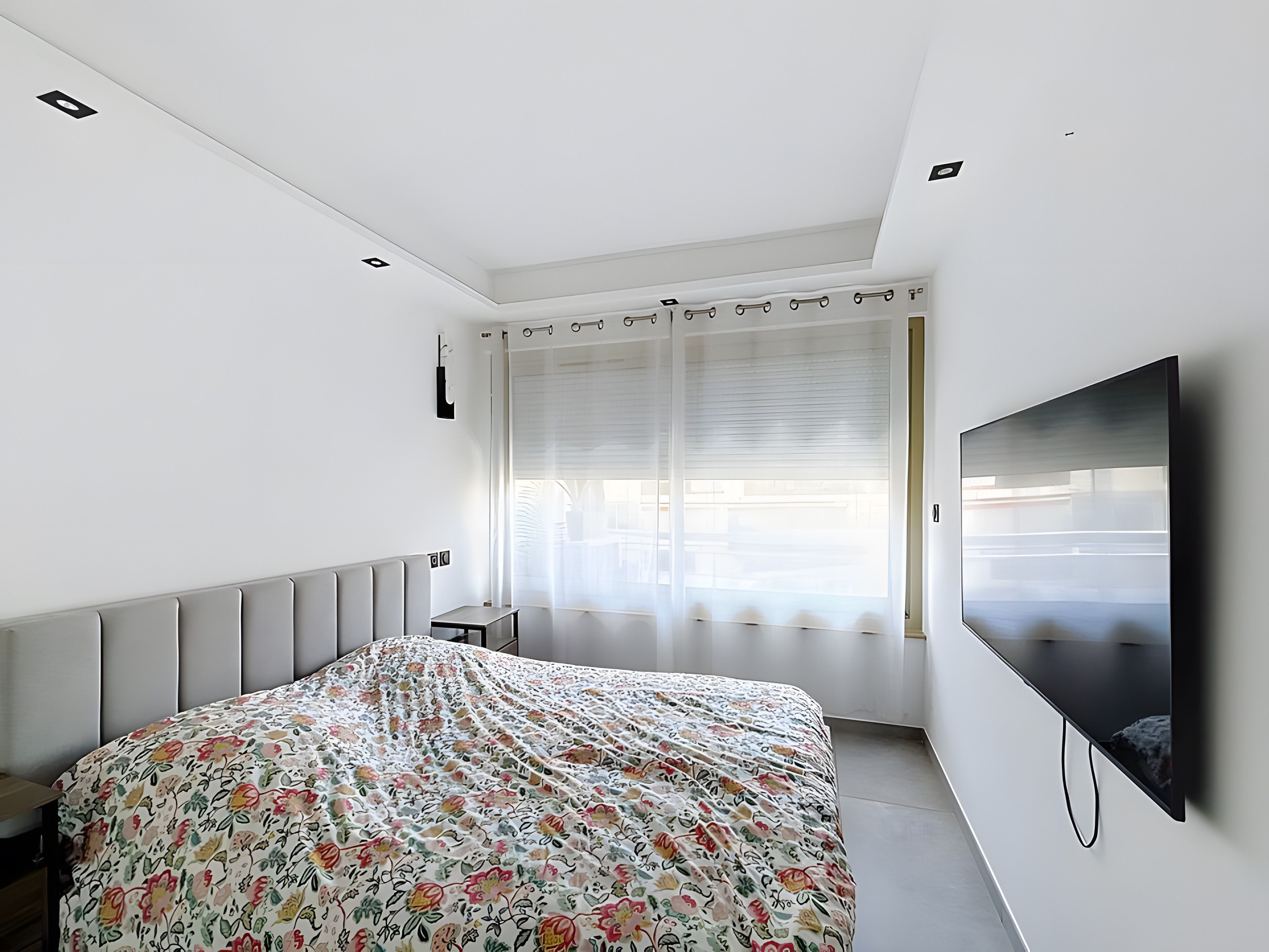 A bedroom with a bed, a TV, a window with blinds, and a radiator.
