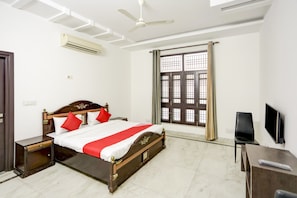 Une chambre d’hôtel comprenant un lit, des tables de chevet, un bureau, une chaise, un téléviseur, un climatiseur, un ventilateur de plafond et une fenêtre avec des rideaux.
