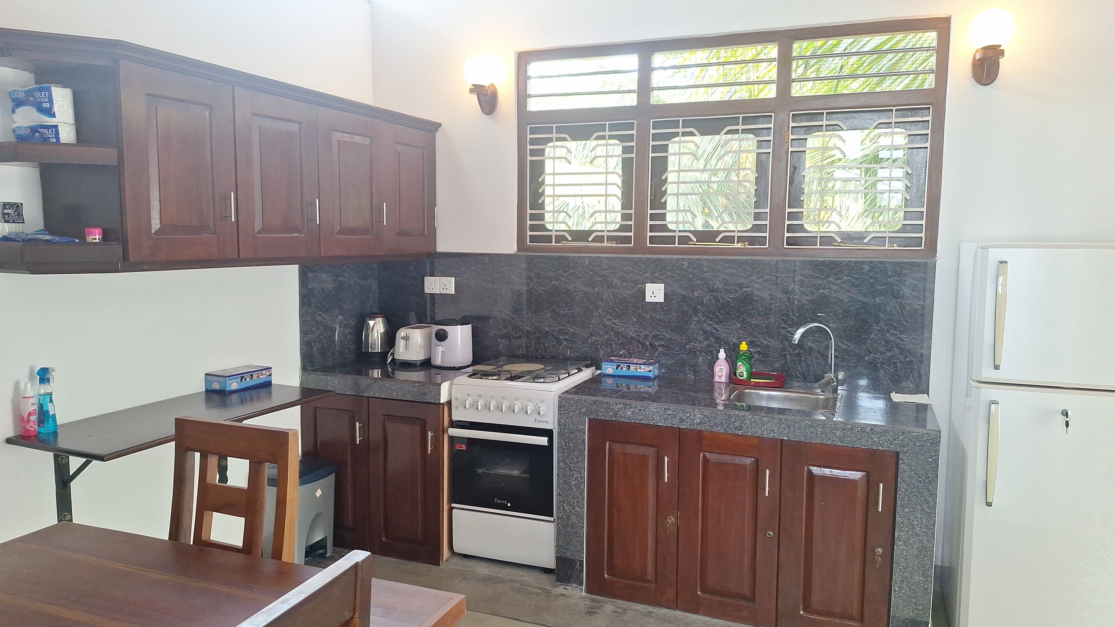 Cocina compacta con estufa a gas, horno y pileta. Hay muebles de madera y una ventana con vista a la vegetación.