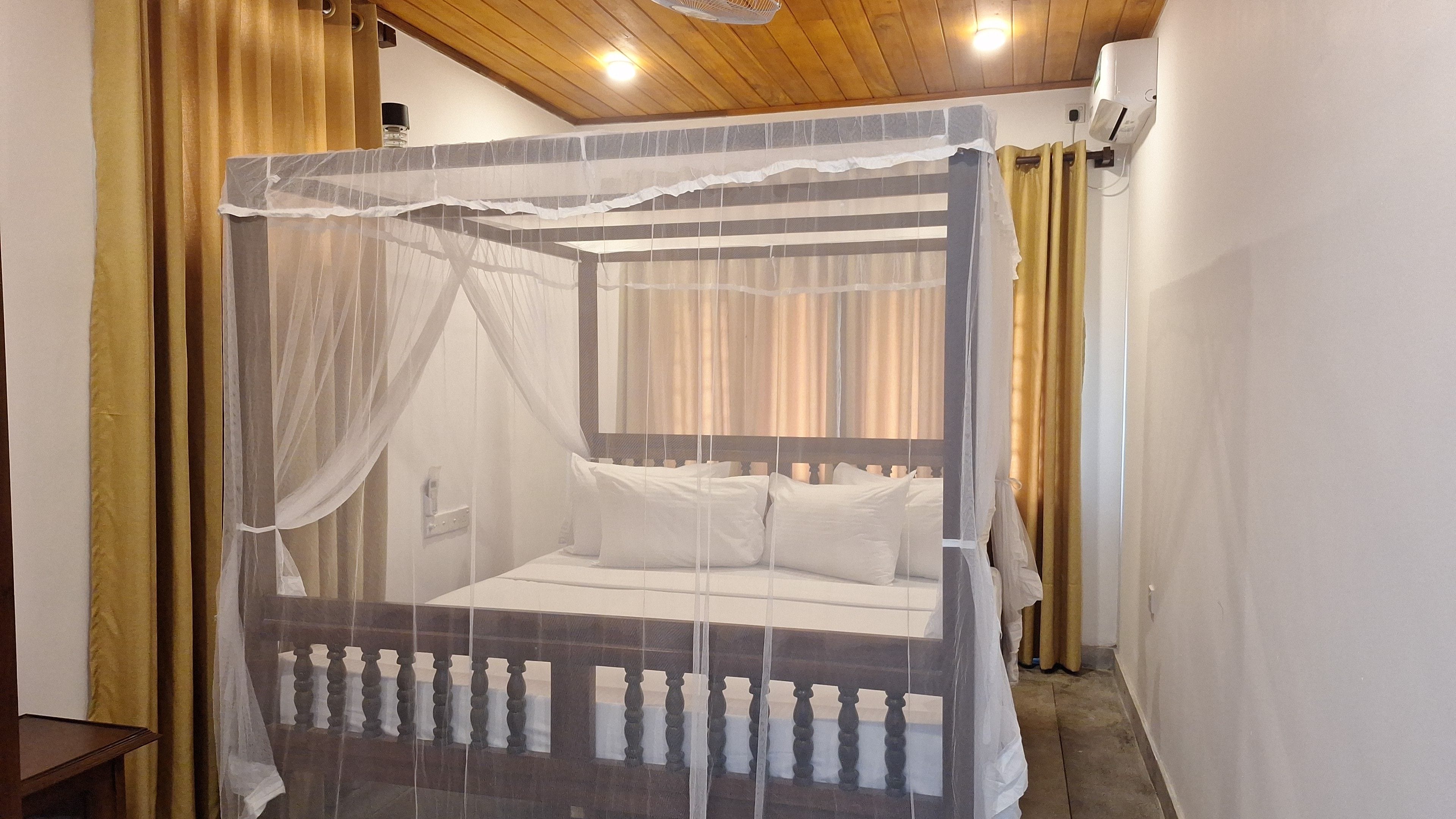 Una cama con dosel sobre pilares, con estructura de madera y ropa de cama blanca, ventilador de techo y aire acondicionado de pared.