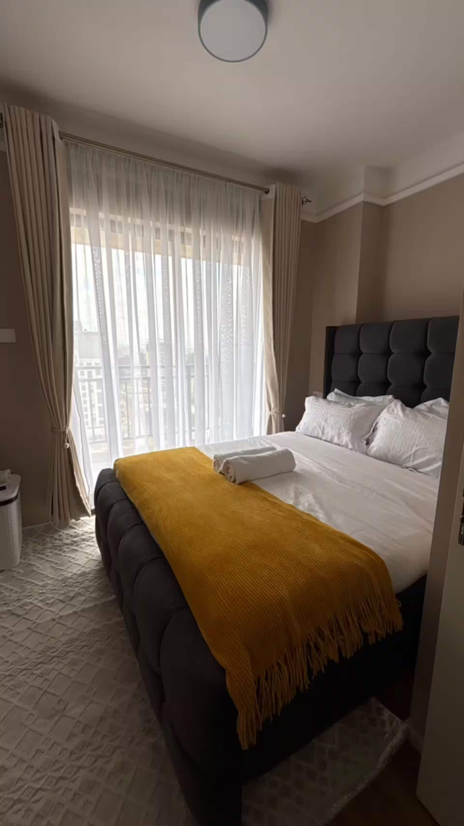 Habitación de hotel con cama, mesita de noche, ventana con cortinas y vista a edificios.