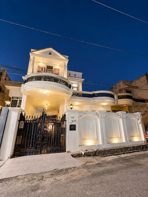 Una casa de varios pisos con balcón, entrada protegida por un portón y fachada bien iluminada.