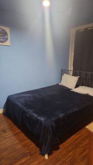 A single bed with a dark blue bedspread and two pillows, a wooden floor, a window with a curtain, and a wall with a circular decoration. - Luxurious Condo Rental with Private Room in Mississauga (Mississauga)