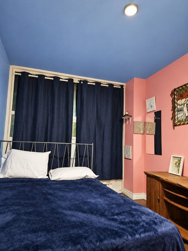 Un dormitori amb llit, cortines, una tauleta de nit i quadres a la paret.