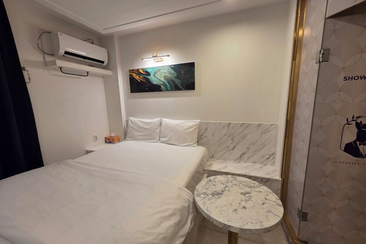 A hotel room with a bed, a small round table, an air conditioning unit, a painting on the wall, and a shower area with a patterned door.