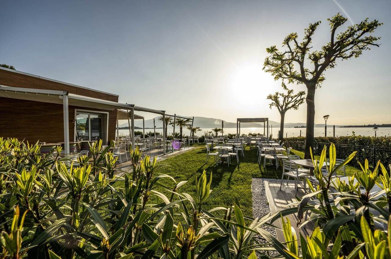 An outdoor dining area with tables and chairs, surrounded by greenery and a view of the sea.