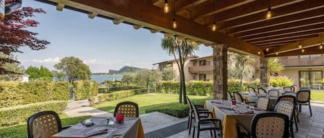 Un área de comedor al aire libre con mesas y sillas, vista al mar y un edificio de piedra al fondo.