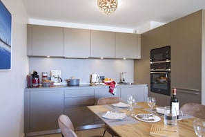 A modern kitchen with grey cabinets, a built-in oven, and a dining area with a wooden table set for two. - R Sidence Phoenix B13 3p6p (Huez)