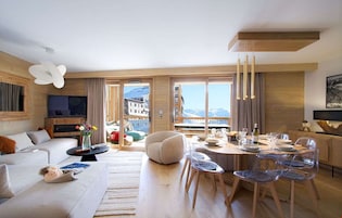 A modern living room with a sofa, coffee table, and dining area with chairs. There is a large window with a view of buildings and mountains.