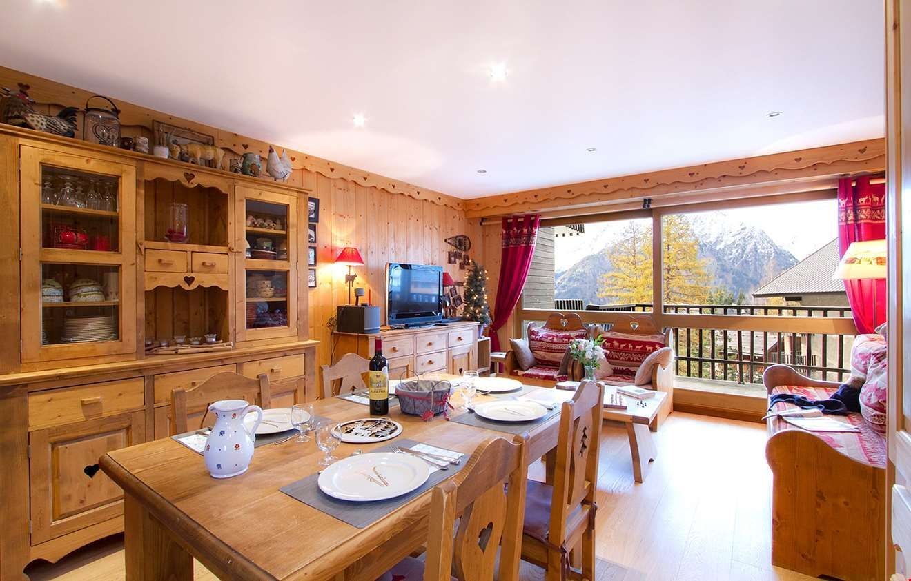 Un comedor de madera con una mesa dispuesta para dos personas, un televisor y vistas a montañas a través de amplios ventanales.