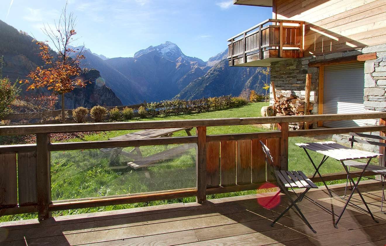 A wooden deck with a view of mountains, a stone wall, and a balcony with a wooden railing.