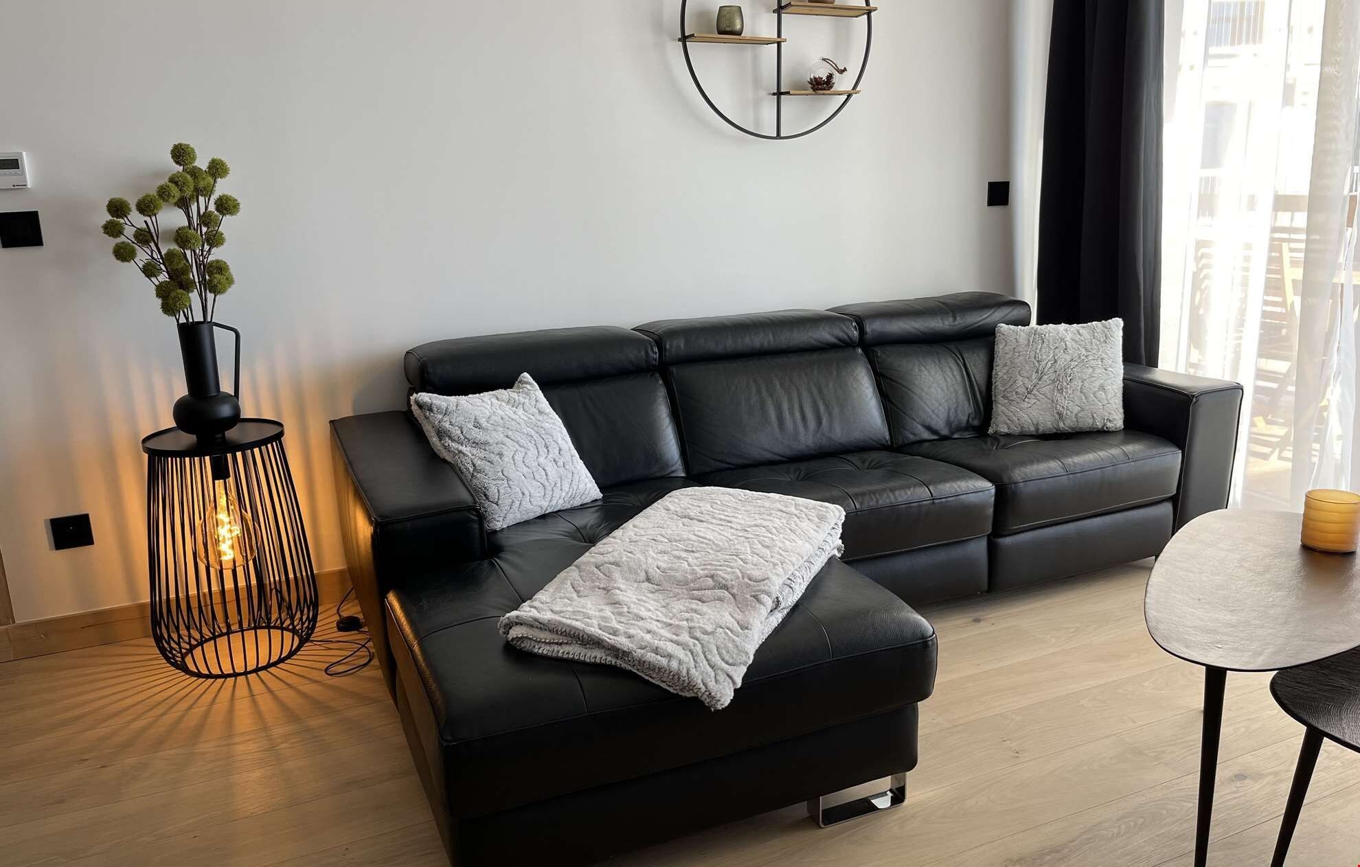 A modern living room with a black leather sofa, two cushions, a side table, a lamp, and a wall-mounted shelf.