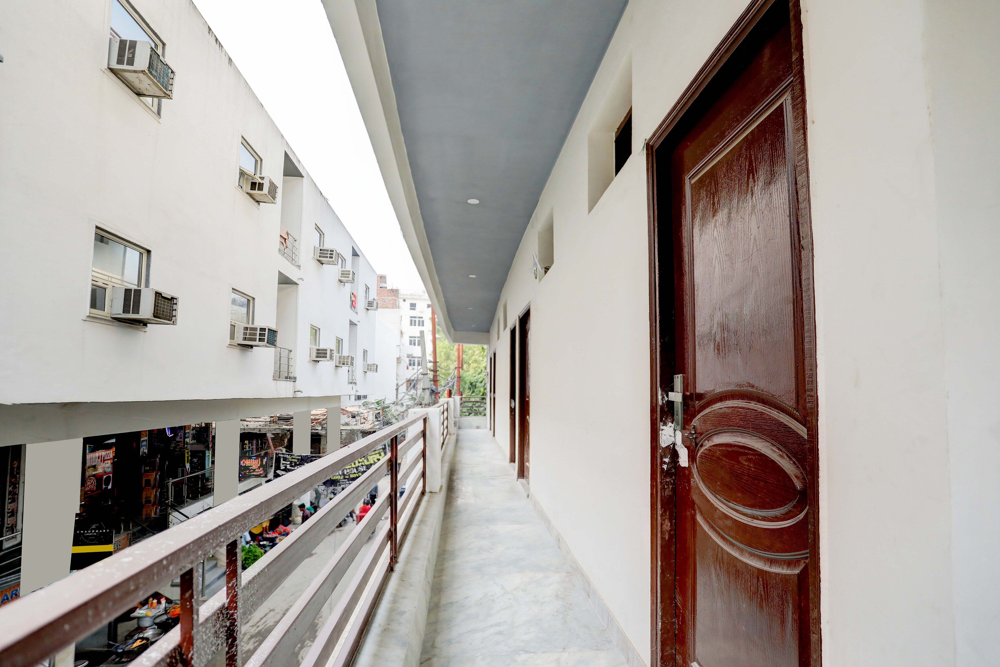 Un couloir avec une porte en bois sur la droite, un balcon avec des balustrades sur la gauche, et plusieurs unités de climatisation fixées aux murs.