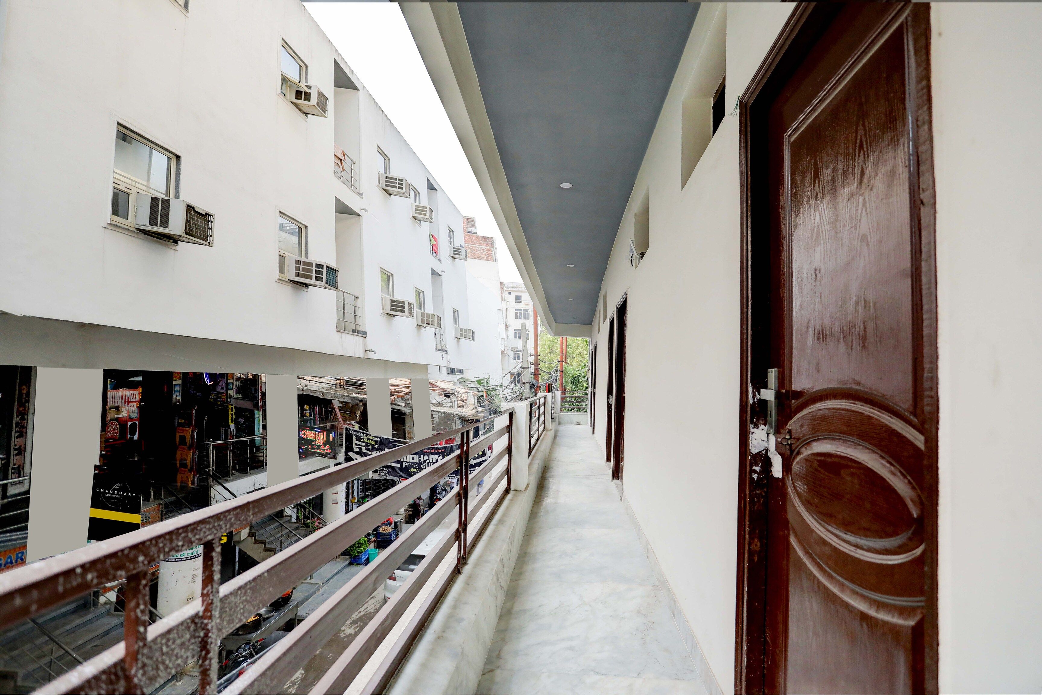 Un pasillo con una puerta de madera a la derecha, un balcón con barandillas y un edificio con múltiples ventanas y unidades de aire acondicionado.