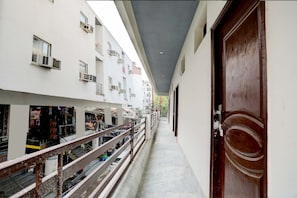 A corridor with a wooden door on the right, a balcony with railings, and a building with multiple windows and air conditioning units.
