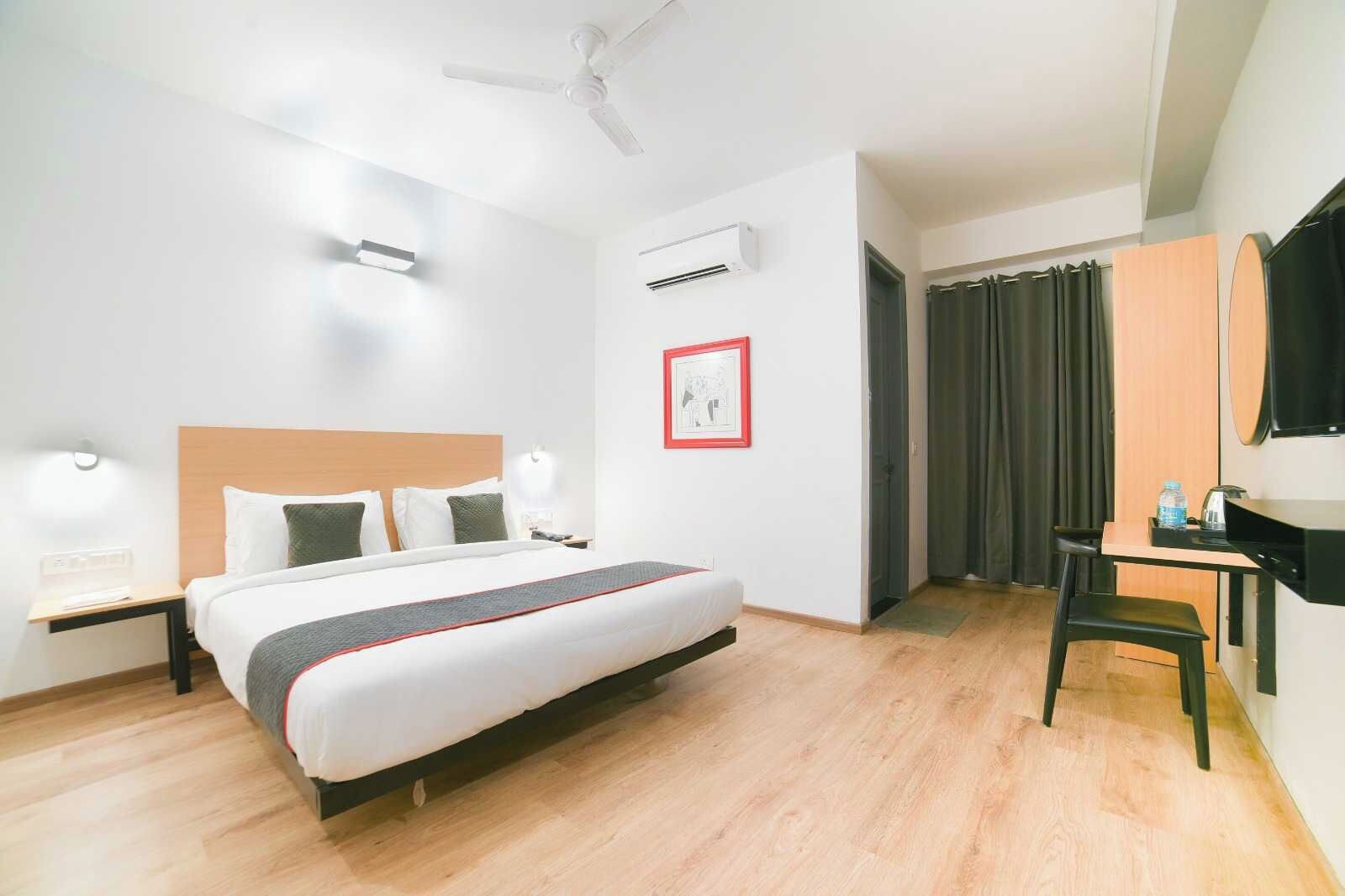 A hotel room with a bed, bedside tables, a desk, a chair, a ceiling fan, an air conditioning unit, a framed picture, and a window with curtains.