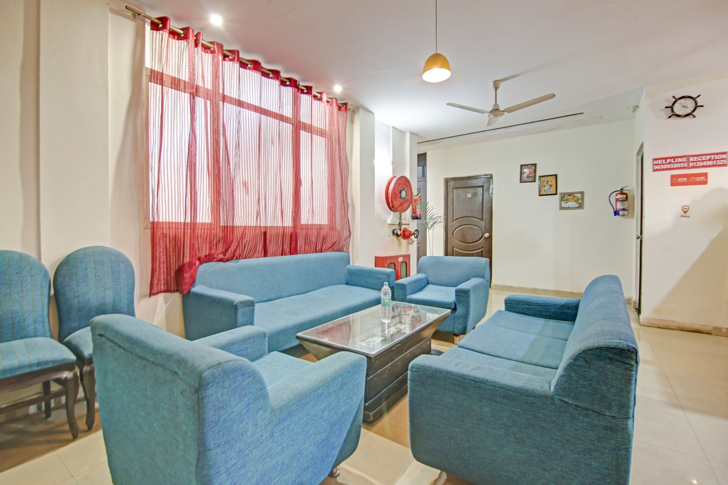 Une chambre d’hôtel avec un canapé bleu, une table basse en verre, un ventilateur de plafond et un rideau rouge.