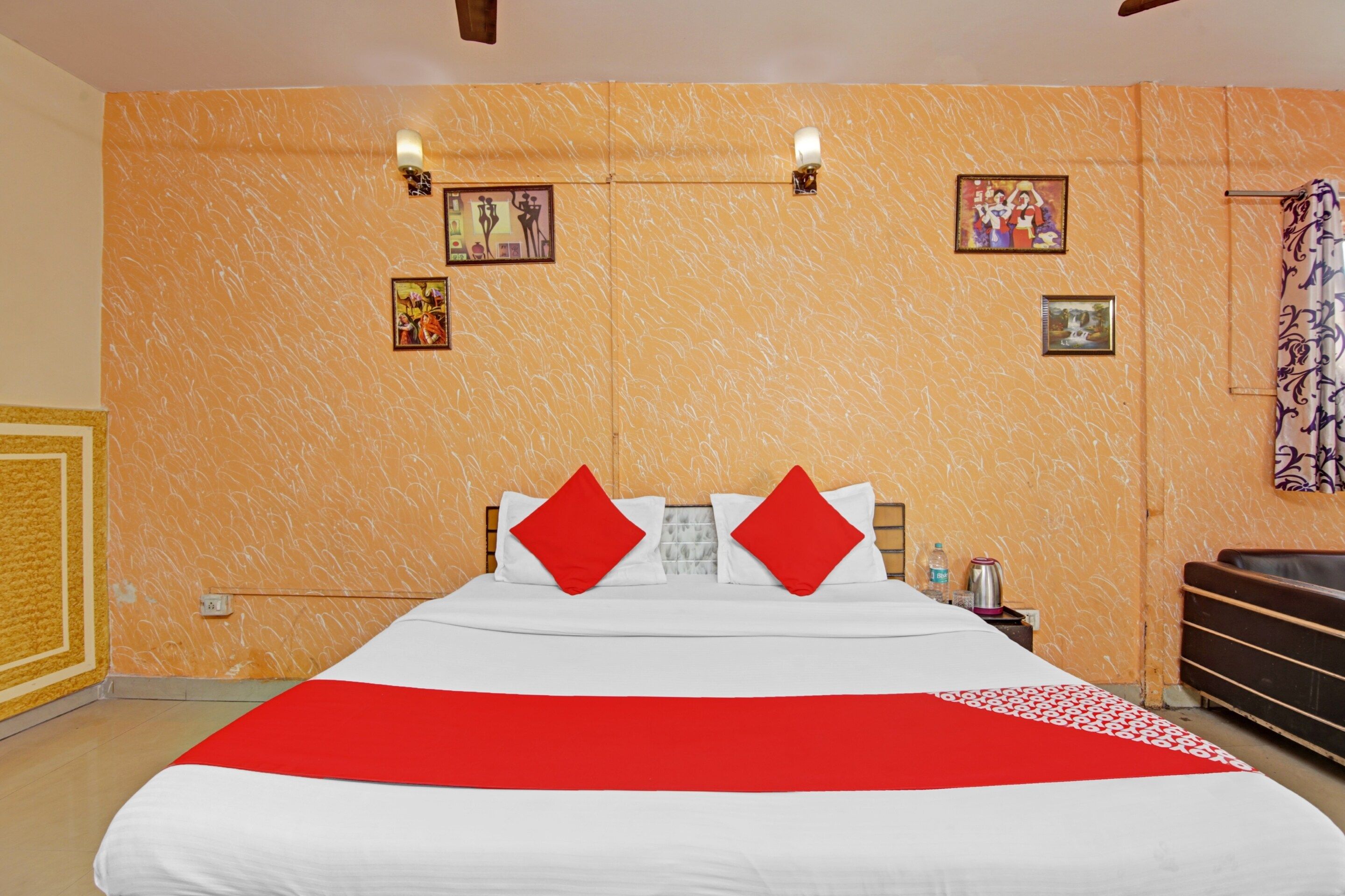Une chambre d’hôtel avec un grand lit, deux oreillers rouges, une couvre-lit blanche et une couverture rayée rouge et blanche. On y trouve des lampes fixées au mur, une petite table avec une bouteille d’eau et une bouilloire, ainsi qu’un rideau à motifs floraux.