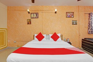 Habitación de hotel con una cama grande, dos almohadas rojas, una colcha blanca y una manta a rayas rojas y blancas. Hay luces fijadas en la pared, una mesita con una botella de agua y una tetera, y una cortina con estampado floral.