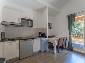 Cocina compacta con microondas, cafetera y lavavajillas. Hay una zona de comedor con mesa y sillas, y una ventana con cortinas.