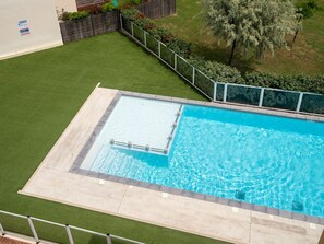 Piscina retangular com revestimento em azulejo, cercada por uma grade de metal e uma área gramada com uma árvore.