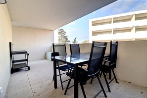 Uma varanda com mesa de vidro e quatro cadeiras, um carrinho pequeno e uma parede branca.