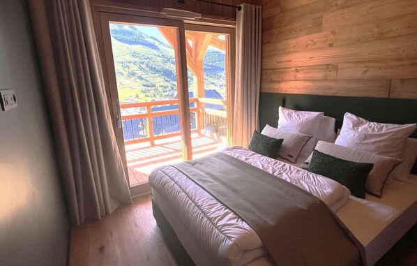 A bedroom with a bed, pillows, a window with curtains, and a view of a balcony. - Chalet des 3 Ours 11p (Les Deux Alpes)
