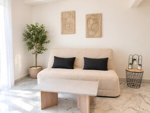 A modern living room with a beige sofa, two black cushions, a wooden coffee table, a potted plant, and a wall-mounted plant stand.