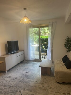 A living room with a flat-screen TV on a white cabinet, a beige sofa with black cushions, a small wooden table, and a hanging light fixture.